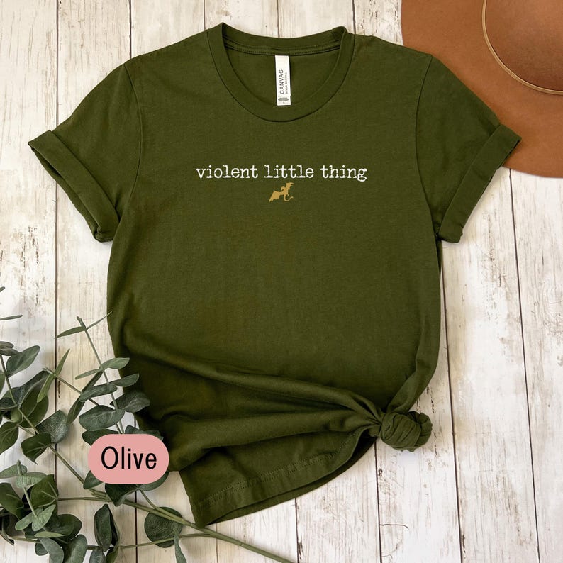 Puede incluir: Camiseta verde oliva con mangas enrolladas y dobladillo anudado. La camiseta presenta el texto "violent little thing" en una fuente serif, con un peque&ntilde;o gr&aacute;fico de perro. La palabra "Olive" est&aacute; en un &oacute;valo rosa.