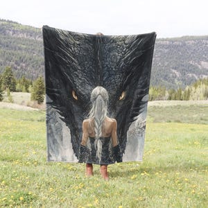 May include: A large, rectangular blanket featuring a detailed illustration of a dragon's head with glowing yellow eyes and a woman with long, silver hair. The blanket is held up in a field of yellow flowers with a mountain backdrop.