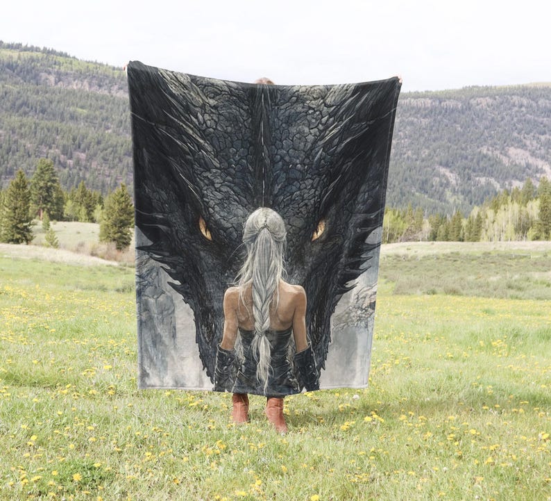 May include: A large, rectangular blanket featuring a detailed illustration of a dragon's head with glowing yellow eyes and a woman with long, silver hair. The blanket is held up in a field of yellow flowers with a mountain backdrop.