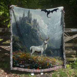 May include: A woven tapestry blanket featuring a fantasy landscape with a white stag in a field of flowers, a castle on a mountain, and a black dragon. The blanket has a fringed edge and is held up outdoors.