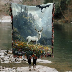 May include: A woven throw blanket featuring a fantasy landscape with a white deer, a castle, and a dragon. The blanket has a colorful floral foreground and a fringed edge with red and brown accents. The background shows a mountain range and a cloudy sky.