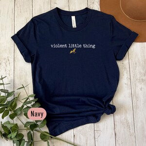 Puede incluir: Camiseta azul marino con el texto blanco "violent little thing" y una peque&ntilde;a ilustraci&oacute;n dorada de un perro. Las mangas est&aacute;n enrolladas y la parte inferior est&aacute; anudada. La palabra "Navy" se muestra en una etiqueta en la esquina inferior izquierda.