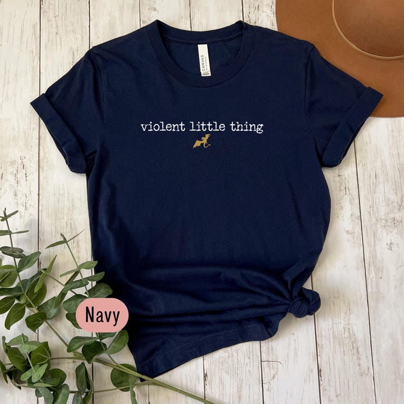 Puede incluir: Camiseta azul marino con el texto blanco "violent little thing" y una peque&ntilde;a ilustraci&oacute;n dorada de un perro. Las mangas est&aacute;n enrolladas y la parte inferior est&aacute; anudada. La palabra "Navy" se muestra en una etiqueta en la esquina inferior izquierda.