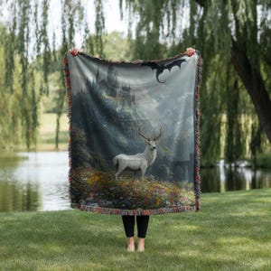 May include: A woven throw blanket featuring a fantasy landscape with a white deer, a castle, and a dragon. The blanket has a fringed edge and is held up against a backdrop of a lake and trees.