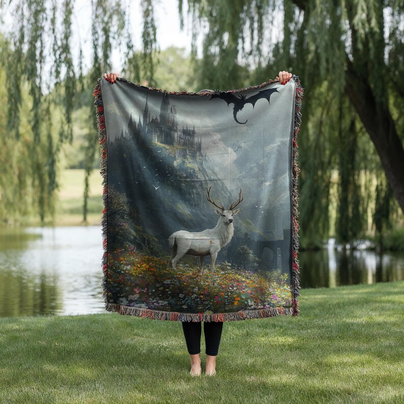 May include: A woven throw blanket featuring a fantasy landscape with a white deer, a castle, and a dragon. The blanket has a fringed edge and is held up against a backdrop of a lake and trees.