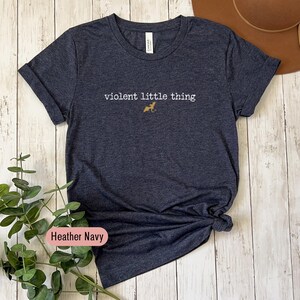 Puede incluir: Camiseta azul marino jaspeado con la frase "violent little thing" y un peque&ntilde;o gr&aacute;fico de perro dorado. La camiseta tiene cuello redondo y est&aacute; hecha de un tejido suave.