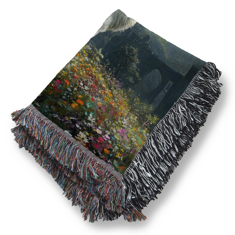 May include: Woven throw blanket featuring a landscape painting with colorful flowers and a dark background. The blanket has a fringed edge with a mix of colors and black and white. The artwork depicts a natural scene.