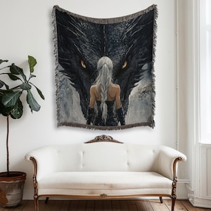 May include: A woven tapestry featuring a fantasy scene with a woman facing a large black dragon. The dragon's eyes are a bright yellow. The tapestry has a fringed border and is displayed on a wall above a white sofa.