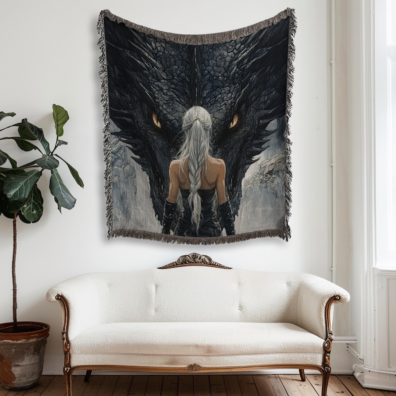 May include: A woven tapestry featuring a fantasy scene with a woman facing a large black dragon. The dragon's eyes are a bright yellow. The tapestry has a fringed border and is displayed on a wall above a white sofa.