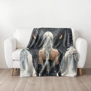 May include: A throw blanket featuring a fantasy art design. The artwork depicts a woman with long, silver hair facing a large black dragon. The dragon's eyes are a bright orange. The woman is wearing a black outfit. The blanket is draped over a white couch.