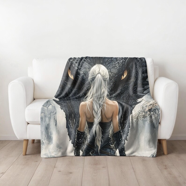 May include: A throw blanket featuring a fantasy art design. The artwork depicts a woman with long, silver hair facing a large black dragon. The dragon's eyes are a bright orange. The woman is wearing a black outfit. The blanket is draped over a white couch.