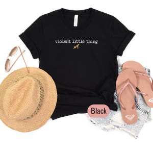 Puede incluir: Camiseta negra con el texto blanco "violent little thing" y un peque&ntilde;o gr&aacute;fico de dinosaurio dorado. La camiseta se combina con un sombrero de paja, gafas de sol y chanclas rosas.