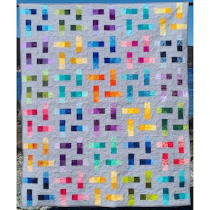 Könnte beinhalten: Eine handgefertigte Steppdecke mit grauem Hintergrund und einem farbenfrohen geometrischen Muster. Das Muster besteht aus rechteckigen Blöcken in verschiedenen Farben, darunter Blau, Grün, Gelb, Orange und Lila. Die Steppdecke wird im Freien ausgestellt.