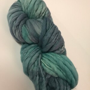 Puede incluir: Un gran ovillo de lana gruesa en tonos turquesa, azul y gris. La lana parece suave y gruesa, ideal para proyectos de tejido o ganchillo. Las variaciones de color crean un efecto jaspeado texturizado.