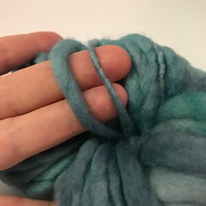 Puede incluir: Primer plano de una mano sosteniendo un bucle de hilo grueso de color verde azulado. El hilo tiene un aspecto suave y texturizado, con variaciones de color de verde azulado claro a oscuro. El hilo se utiliza probablemente para tejer u otras manualidades con fibra.