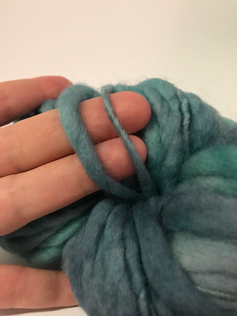 Puede incluir: Primer plano de una mano sosteniendo un bucle de hilo grueso de color verde azulado. El hilo tiene un aspecto suave y texturizado, con variaciones de color de verde azulado claro a oscuro. El hilo se utiliza probablemente para tejer u otras manualidades con fibra.