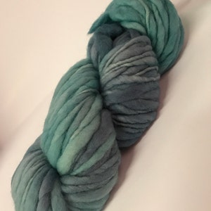 Puede incluir: Un ovillo de lana gruesa te&ntilde;ida a mano en tonos turquesa y gris. La lana gruesa est&aacute; fuertemente torcida, mostrando variaciones de color y textura. Una etiqueta est&aacute; adherida a la parte superior del ovillo. Ideal para proyectos de tejido o ganchillo.