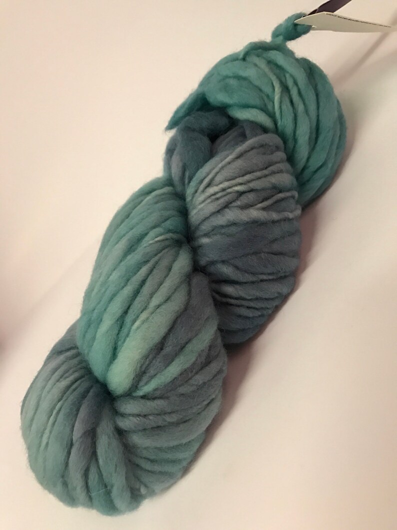Puede incluir: Un ovillo de lana gruesa te&ntilde;ida a mano en tonos turquesa y gris. La lana gruesa est&aacute; fuertemente torcida, mostrando variaciones de color y textura. Una etiqueta est&aacute; adherida a la parte superior del ovillo. Ideal para proyectos de tejido o ganchillo.