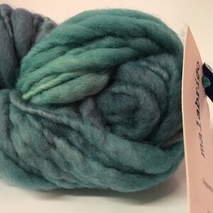 Puede incluir: Un gran ovillo de lana verde azulado y azul verdoso. La lana es gruesa y voluminosa, con un aspecto suave y texturizado. Una etiqueta con la marca "malabrigo" est&aacute; adherida a la lana. La lana es adecuada para proyectos de tejido o ganchillo.