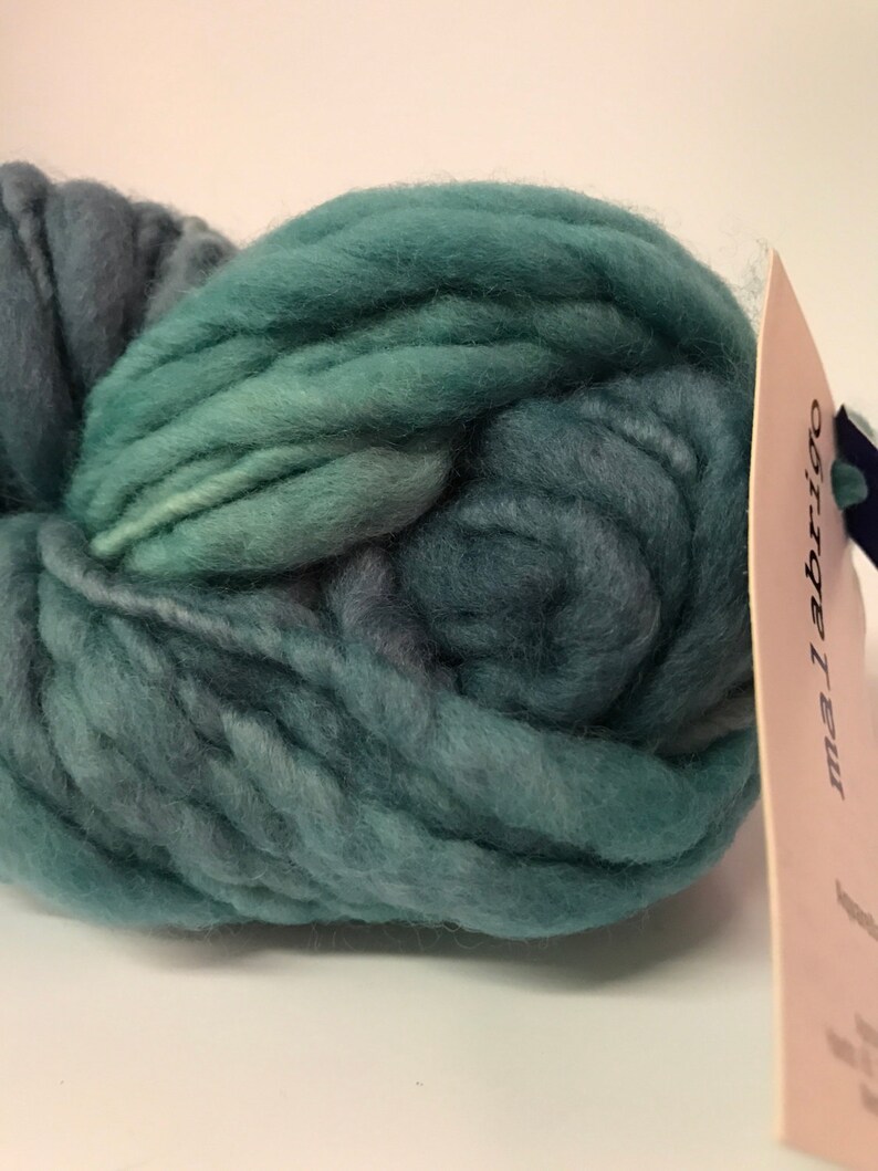Puede incluir: Un gran ovillo de lana verde azulado y azul verdoso. La lana es gruesa y voluminosa, con un aspecto suave y texturizado. Una etiqueta con la marca "malabrigo" est&aacute; adherida a la lana. La lana es adecuada para proyectos de tejido o ganchillo.
