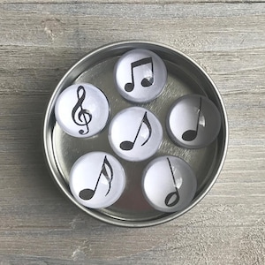 May include: A set of six round glass magnets with black musical note and treble clef designs on a white background. The magnets are in a silver tin.