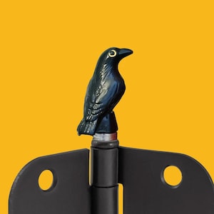 May include: A black crow-shaped decorative hinge. The crow has a detailed design with a yellow eye. The hinge is black with two circular holes. The background is a solid yellow.