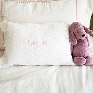 May include: A white pillow with a pink embroidered design and the text "Just Jill" on a white bed with a pink plush toy dog.