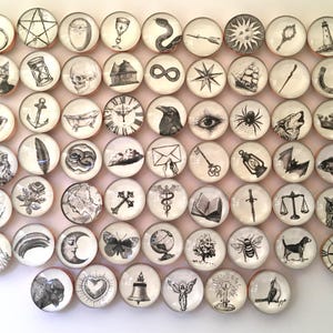 May include: A collection of round, white and beige decorative buttons with black and white illustrations. The illustrations include a ring, a pentagram, a cat, an hourglass, a skull, a house, an infinity symbol, a compass, a ship, a feather, a rose, and a butterfly.