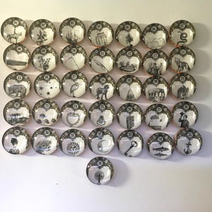 May include: A collection of thirty-six round, decorative cabinet knobs. Each knob features a black and white illustration with a gold-colored border and a number. The illustrations depict various scenes and objects.