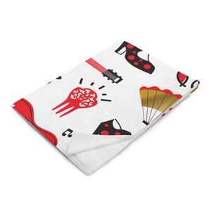 Puede incluir: Manta blanca con un patrón repetido de zapatos de flamenco rojos y negros, una guitarra roja, una castañuela roja, un abanico dorado y notas musicales. La manta está doblada, mostrando la parte inferior blanca.