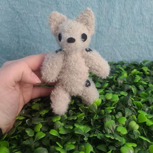 May include: A small, handmade plush toy in the shape of a bear. The bear is light beige with black button eyes and small black accents on its limbs. It has a fluffy texture and is standing on a bed of green leaves.