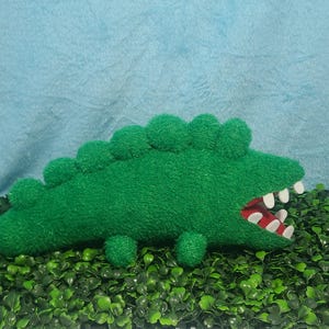 May include: Green plush crocodile toy with a toothy grin and a red mouth. The toy is lying on a bed of green artificial grass.