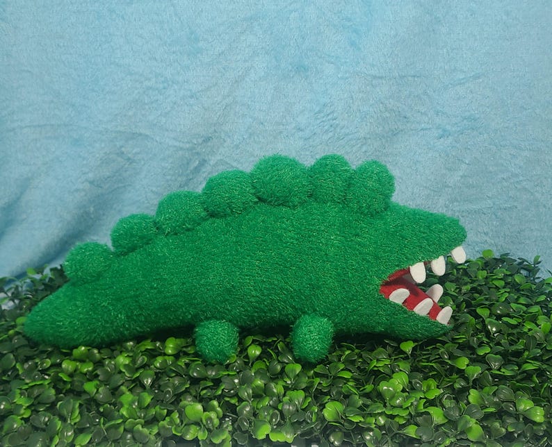 May include: Green plush crocodile toy with a toothy grin and a red mouth. The toy is lying on a bed of green artificial grass.