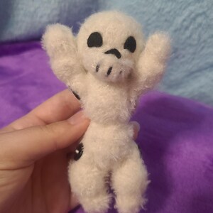May include: A small, handmade plush toy in the shape of a skull. The white plush figure has black eyes and a mouth, with arms raised. The toy is held against a purple background.