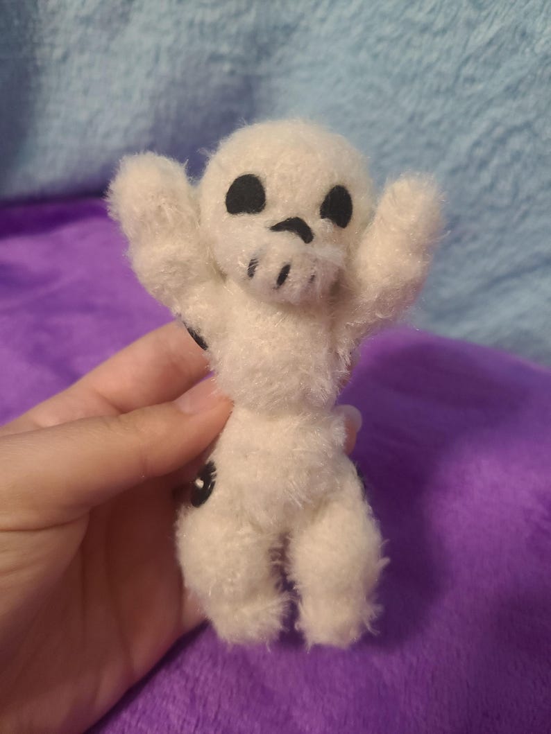 May include: A small, handmade plush toy in the shape of a skull. The white plush figure has black eyes and a mouth, with arms raised. The toy is held against a purple background.
