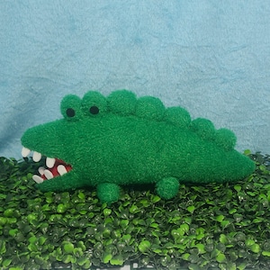 May include: A green plush alligator toy with white teeth. The alligator is lying on a bed of green artificial grass.
