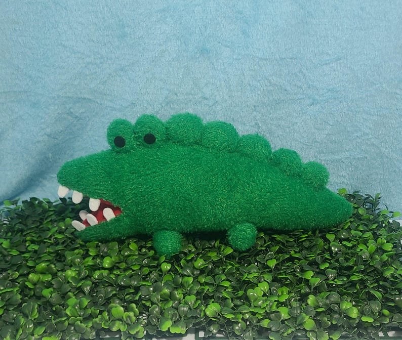 May include: A green plush alligator toy with white teeth. The alligator is lying on a bed of green artificial grass.