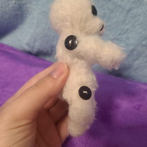 May include: A small, handmade white plush toy with black button eyes and two black buttons on the body. The toy has a simple, rounded shape and a fuzzy texture, suggesting it is made of yarn or a similar material. The toy is held in a hand.