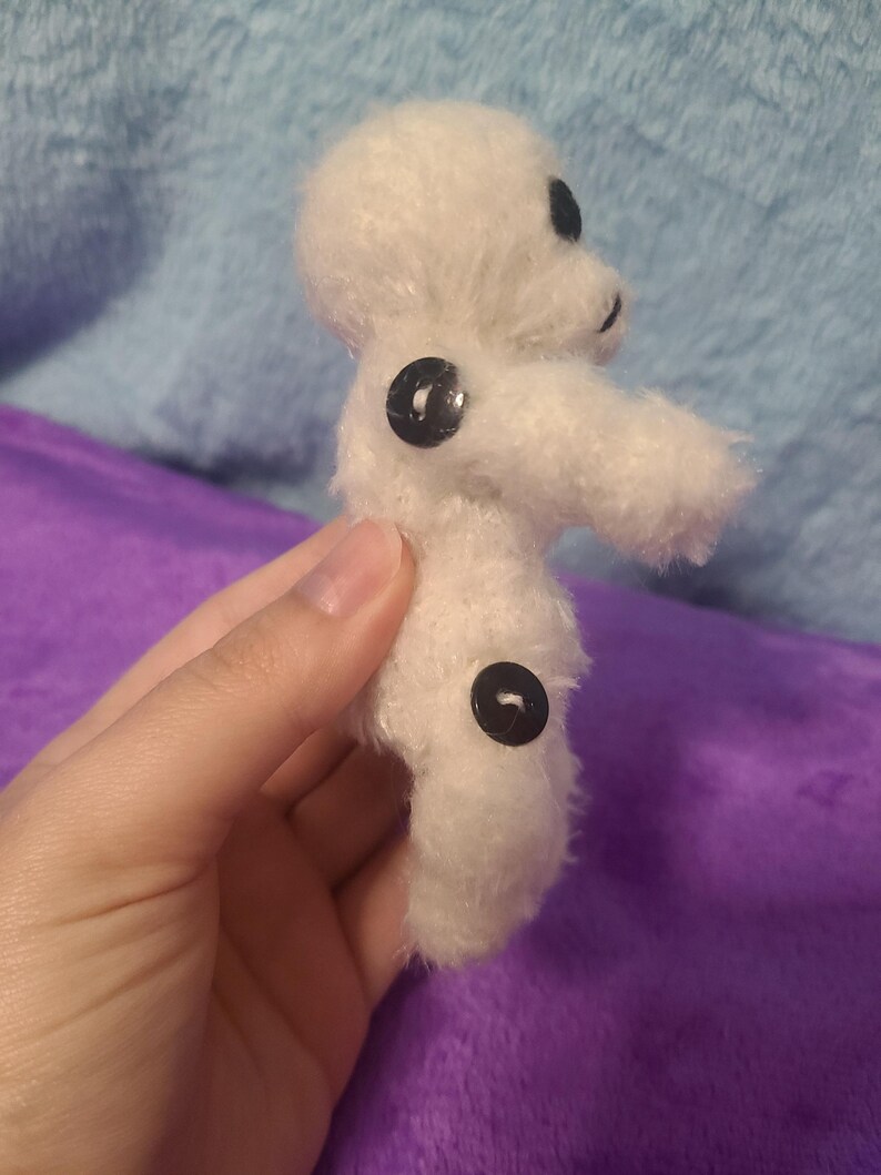 May include: A small, handmade white plush toy with black button eyes and two black buttons on the body. The toy has a simple, rounded shape and a fuzzy texture, suggesting it is made of yarn or a similar material. The toy is held in a hand.