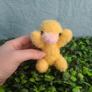 May include: A small, handmade yellow plush duck with a pink beak and black eyes. The duck has outstretched arms and a fluffy texture. The background is a mix of green foliage and a light blue surface.