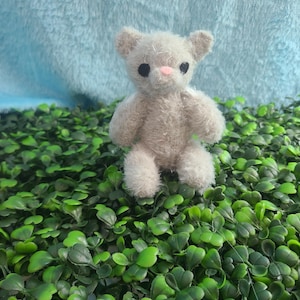 May include: A small, plush teddy bear with light gray fur, black eyes, and a pink nose. The bear is standing on a bed of green leaves against a light blue, textured background. The bear's arms and legs are short and stubby.