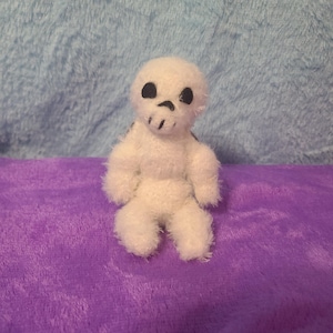 May include: A small, white, fuzzy skull-shaped plush toy with black eyes and a mouth. The toy is sitting on a purple surface with a blue background. The plushie has small arms and legs.