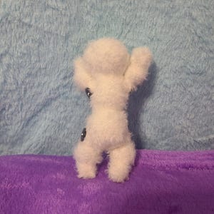 May include: A small, white, handmade felt doll with outstretched arms and two black button eyes. The doll is standing on a purple surface against a light blue background.