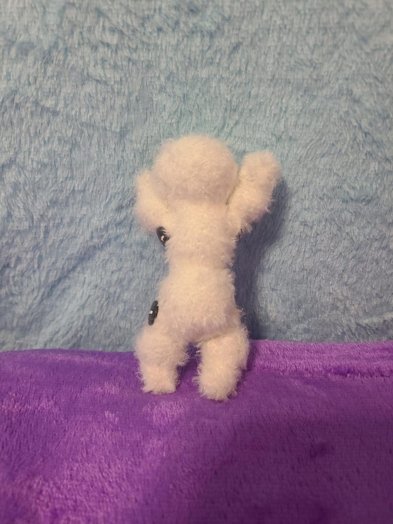 May include: A small, white, handmade felt doll with outstretched arms and two black button eyes. The doll is standing on a purple surface against a light blue background.