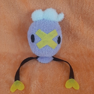 May include: A purple plush toy with a yellow X over its eyes and black arms with yellow heart-shaped hands. The toy has a blue fluffy head.