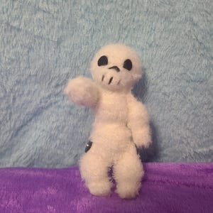 May include: A small, white, fuzzy doll in the shape of a skull. The doll has black eyes and a mouth, and is posed with one arm raised. The doll is on a purple surface with a blue background.