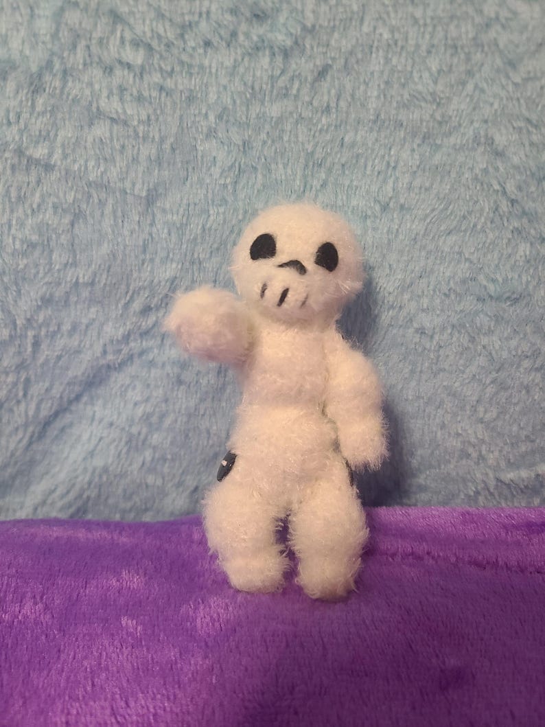May include: A small, white, fuzzy doll in the shape of a skull. The doll has black eyes and a mouth, and is posed with one arm raised. The doll is on a purple surface with a blue background.