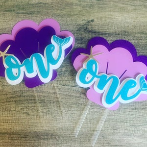 May include: Two purple and pink seashell-shaped cake toppers with the word "one" in turquoise glitter letters. Each topper has a gold glitter starfish and a turquoise glitter mermaid tail.