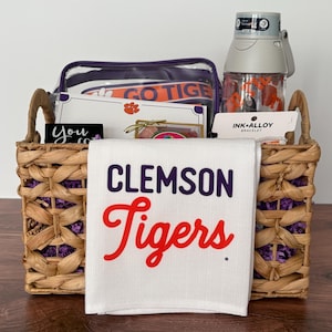 May include: A woven wicker basket filled with purple paper shred, containing a white towel with the text CLEMSON Tigers, a clear plastic water bottle with a grey lid, a clear zippered pouch with the text GO TIGERS, and an Ink+Alloy bracelet.