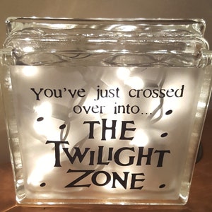 May include: A glass block with a white background and the text "You've just crossed over into... THE TWILIGHT ZONE."  The block is illuminated with white fairy lights.